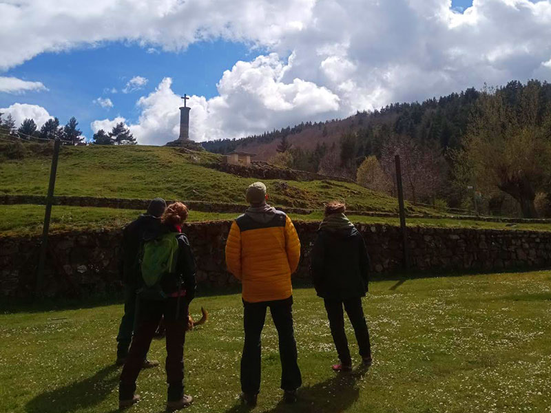 Cuatro personas de espaldas observan una colina verde con una cruz en la cima, bajo un cielo parcialmente nublado y rodeados de árboles.