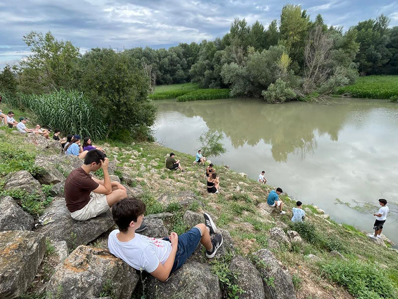 Varias personas están sentadas en una ladera cubierta de hierba y piedras, observando un río tranquilo rodeado de vegetación frondosa bajo un cielo nublado.
