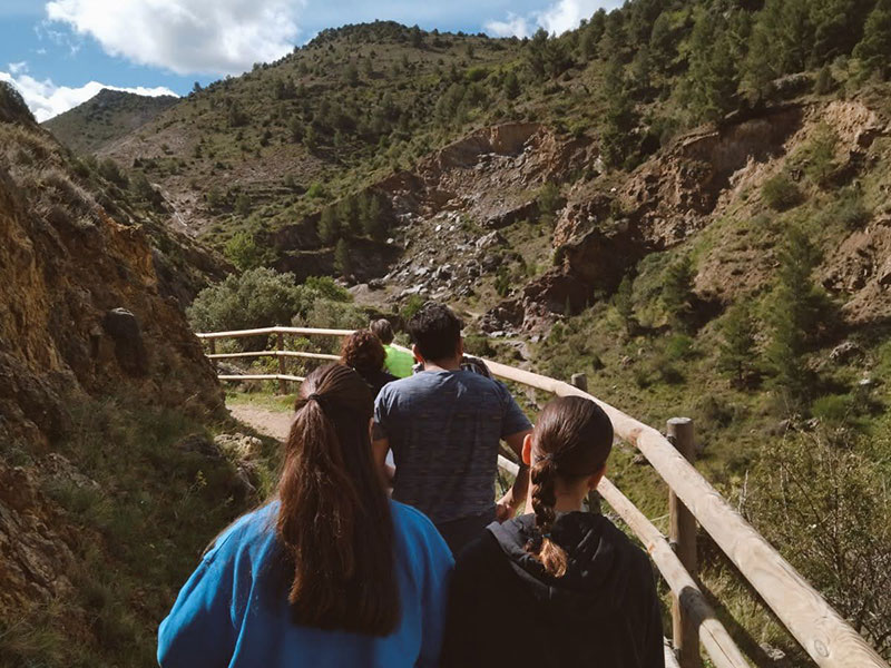 Un grupo de personas camina por un sendero rodeado de montañas verdes y rocosas, con barandillas de madera a los lados.