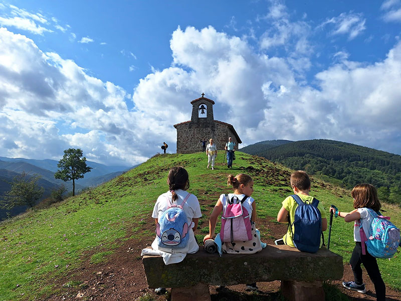 Un grupo de niños con mochilas coloridas descansa en un banco de piedra en una colina verde, mientras observan una pequeña capilla de piedra con campanario bajo un cielo parcialmente nublado. Al fondo, se ven montañas y dos adultos caminando hacia los niños.